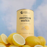 A can of Nutra Naturals Protein Water Lemonade 450g, rich in electrolytes, stands upright against a blue sky with whole and halved fresh lemons placed in the foreground.