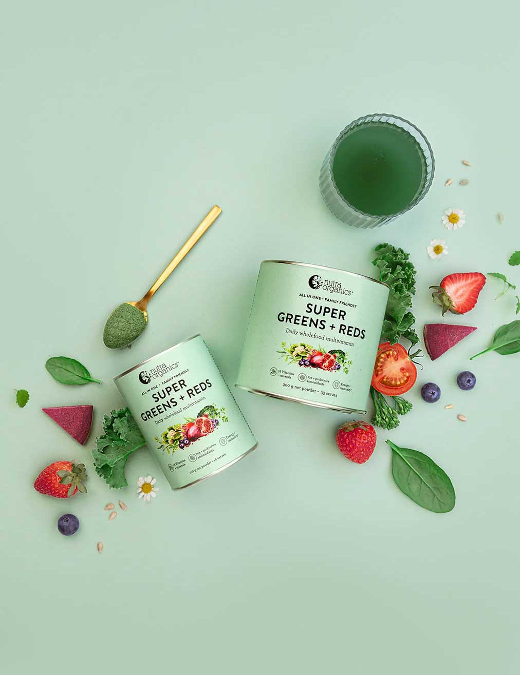 Two containers of Nutra Naturals Super Greens + Reds with a scoop, surrounded by superfoods, leafy greens, strawberries, spinach, blueberries, tomato, and a green drink in a glass, all set on a pale green background.