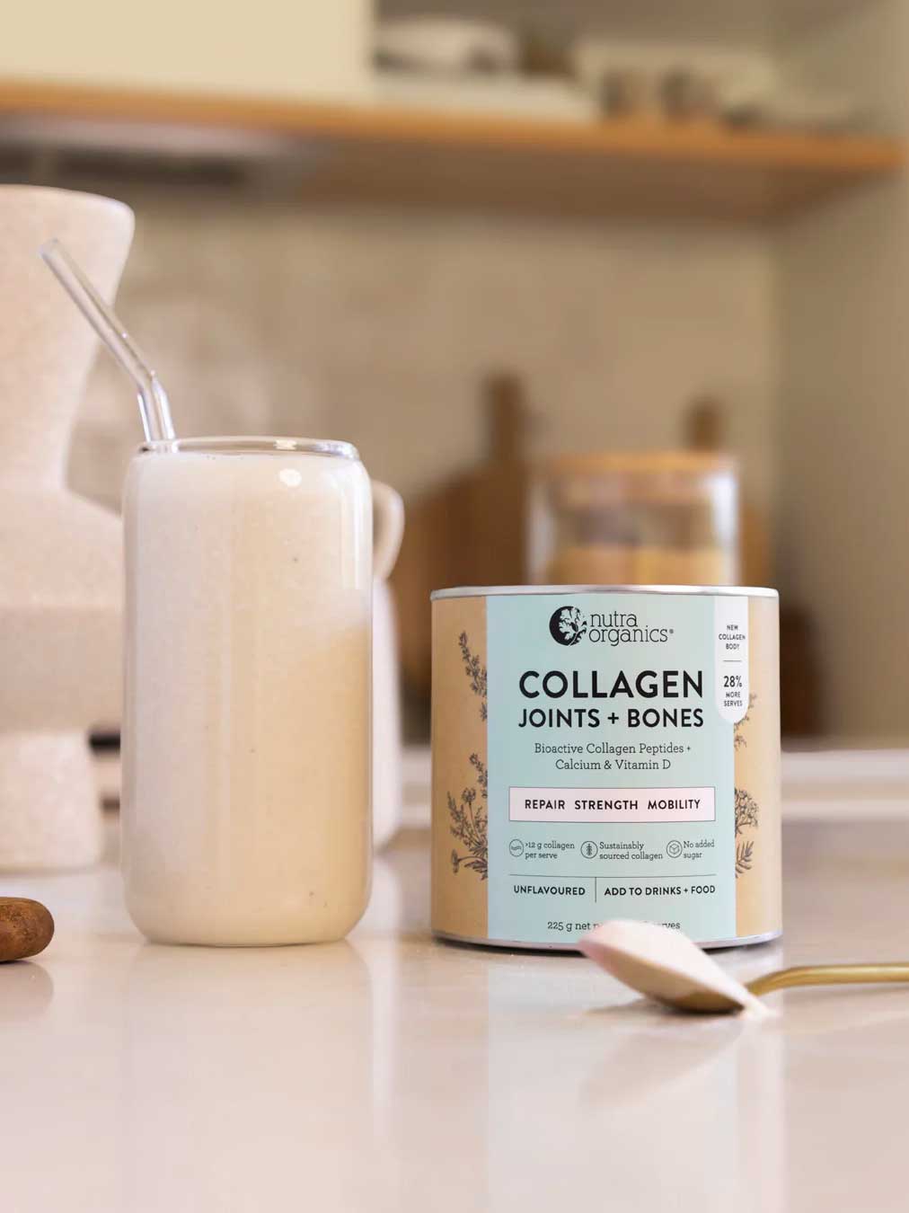 A creamy collagen powder drink with a straw sits next to a canister of Nutra Naturals Collagen Joints + Bones on a kitchen counter, emphasizing bone health support. A spoon lies nearby, with the softly blurred background adding warmth.