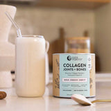 A creamy collagen powder drink with a straw sits next to a canister of Nutra Naturals Collagen Joints + Bones on a kitchen counter, emphasizing bone health support. A spoon lies nearby, with the softly blurred background adding warmth.