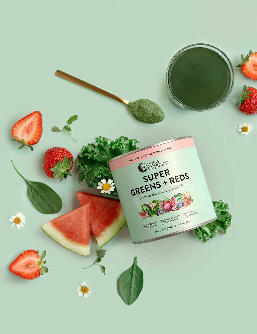 A jar of Nutra Naturals Super Greens + Reds Watermelon Strawberry 300g sits on a light green background, surrounded by strawberries, watermelon slices, spinach, kale, chamomile flowers, and a gold spoon filled with green powder.