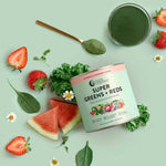 A jar of Nutra Naturals Super Greens + Reds Watermelon Strawberry 300g sits on a light green background, surrounded by strawberries, watermelon slices, spinach, kale, chamomile flowers, and a gold spoon filled with green powder.