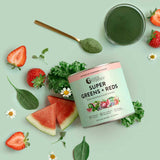 A jar of Nutra Naturals Super Greens + Reds Watermelon Strawberry 300g sits on a light green background, surrounded by strawberries, watermelon slices, spinach, kale, chamomile flowers, and a gold spoon filled with green powder.