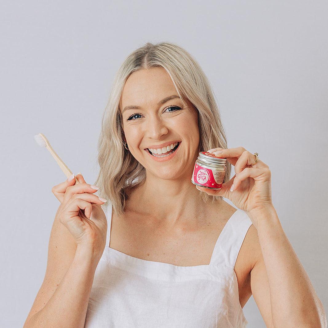 Woman smiling with a toothbrush and Oh Gigi Fruity Hydroxyapatite Tooth Powder