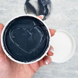 A hand holds an open Elektra cleanser jar towards the camera, showing thick charcoal-grey cream with curved scoop marks and a matching smear on the surface behind.