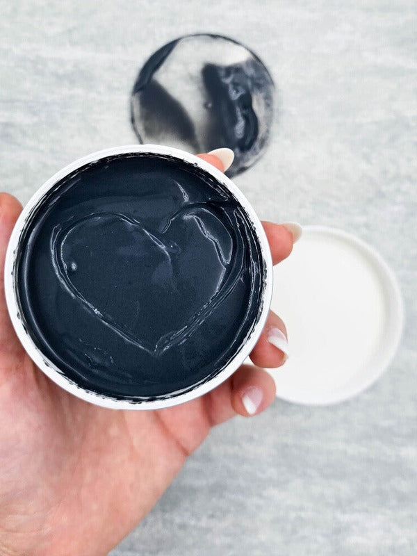 A hand holds an open Elektra cleanser jar towards the camera, showing thick charcoal-grey cream with curved scoop marks and a matching smear on the surface behind.