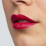 Close-up of smooth skin and full lips wearing Antipodes Moisture Boost Natural Lipstick in a vibrant, glossy plum shade. The text "Oriental Bay Plum" highlights its long-lasting color.