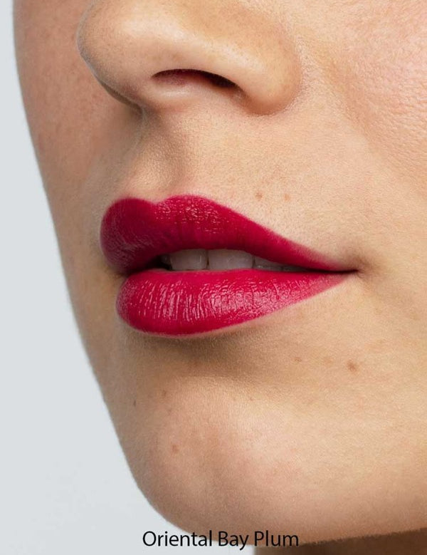 Close-up of smooth skin and full lips wearing Antipodes Moisture Boost Natural Lipstick in a vibrant, glossy plum shade. The text "Oriental Bay Plum" highlights its long-lasting color.