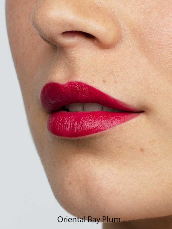 Close-up of smooth skin and full lips wearing Antipodes Moisture Boost Natural Lipstick in a vibrant, glossy plum shade. The text "Oriental Bay Plum" highlights its long-lasting color.
