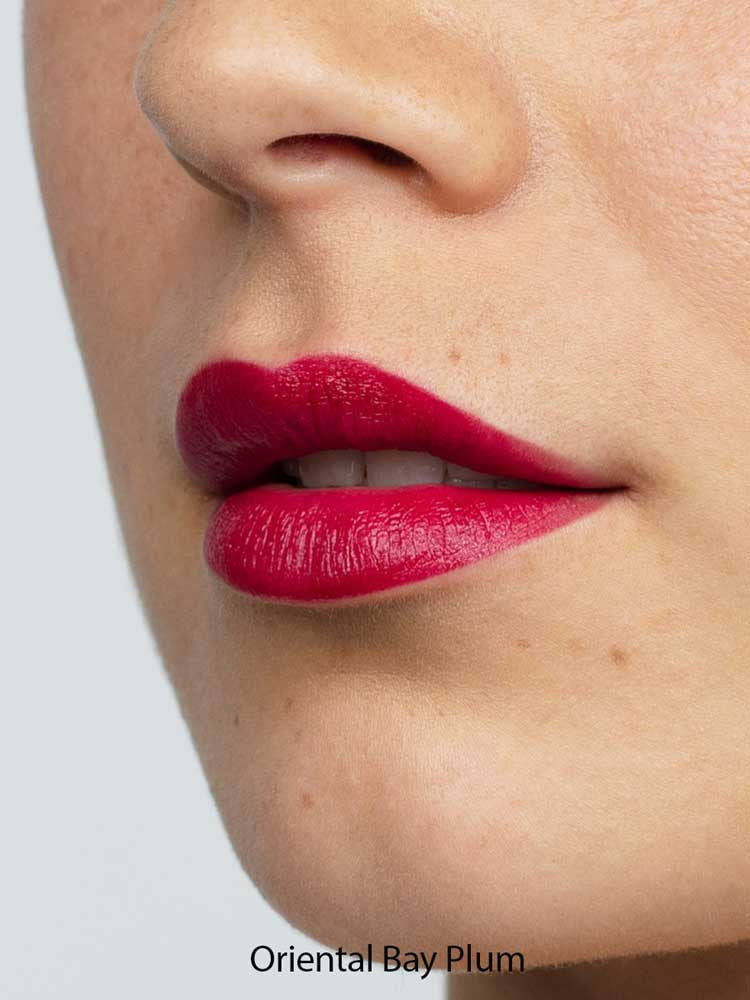 Close-up of smooth skin and full lips wearing Antipodes Moisture Boost Natural Lipstick in a vibrant, glossy plum shade. The text "Oriental Bay Plum" highlights its long-lasting color.