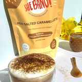 A glass mug of frothy salted caramel latte sits beside a wooden spoon, with a yellow packet of She BANG! Peri Salted Caramel Latte 300g and yellow flowers in the background.