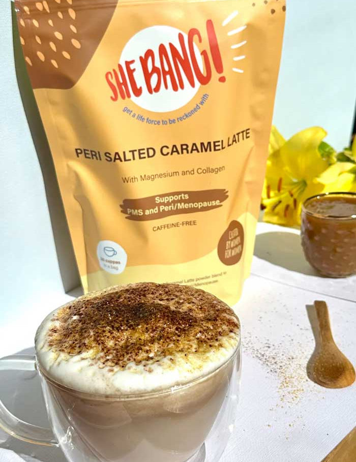 A glass mug of frothy salted caramel latte sits beside a wooden spoon, with a yellow packet of She BANG! Peri Salted Caramel Latte 300g and yellow flowers in the background.