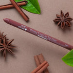 A brown 100% Pure lip liner labelled Chai lies diagonally on a taupe surface among cinnamon sticks, star anise and green leaves. Its rounded tip points left.