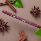 A brown 100% Pure lip liner labelled Chai lies diagonally on a taupe surface among cinnamon sticks, star anise and green leaves. Its rounded tip points left.