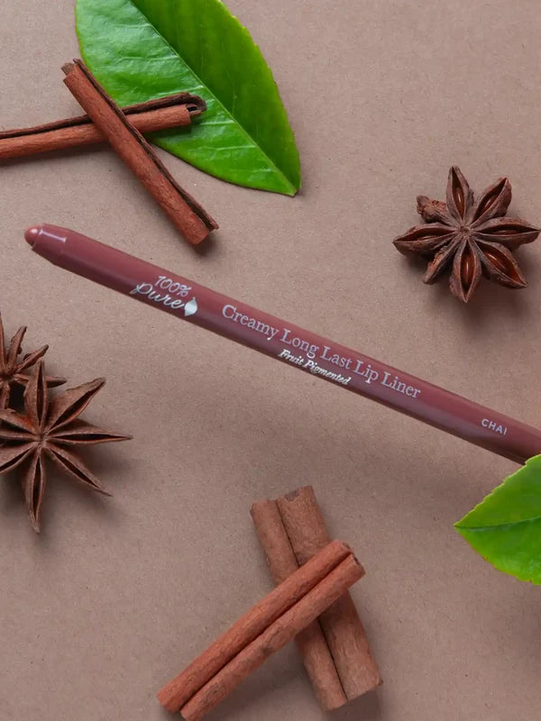 A brown 100% Pure lip liner labelled Chai lies diagonally on a taupe surface among cinnamon sticks, star anise and green leaves. Its rounded tip points left.