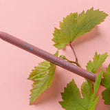 A muted pink-brown 100% Pure lip liner lies diagonally across green vine leaves on a pale pink surface. The barrel is labelled Cognac.