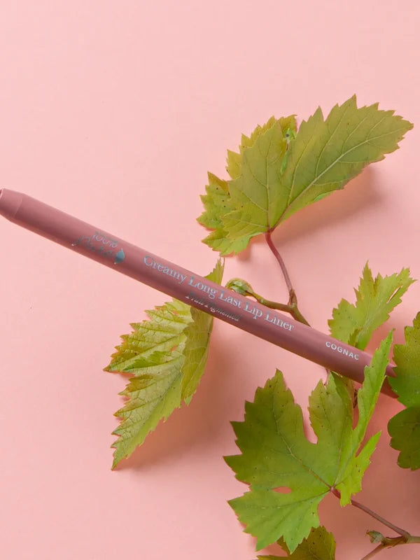 A muted pink-brown 100% Pure lip liner lies diagonally across green vine leaves on a pale pink surface. The barrel is labelled Cognac.