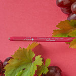 A pink-red 100% Pure lip liner lies horizontally between clusters of purple grapes and green vine leaves on a deep pink surface. Its tip points left, and Pink Brandy is printed on the barrel.