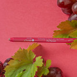 A pink-red 100% Pure lip liner lies horizontally between clusters of purple grapes and green vine leaves on a deep pink surface. Its tip points left, and Pink Brandy is printed on the barrel.