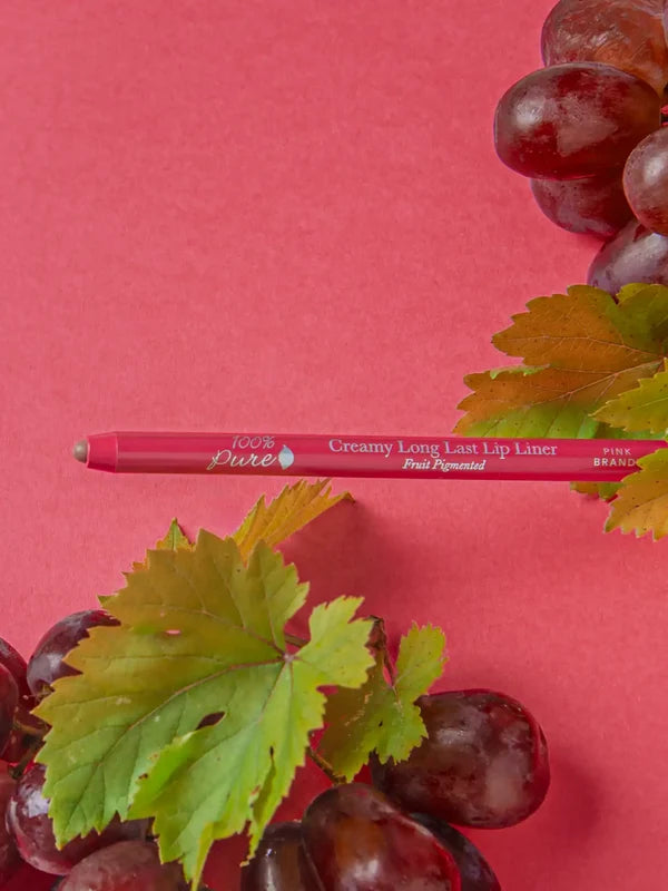 A pink-red 100% Pure lip liner lies horizontally between clusters of purple grapes and green vine leaves on a deep pink surface. Its tip points left, and Pink Brandy is printed on the barrel.