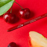 A red 100% Pure lip liner lies diagonally on a bright red surface beside cherries, a green leaf, a dark grape and a pale fruit wedge with red skin. Its exposed tip points toward the lower left.
