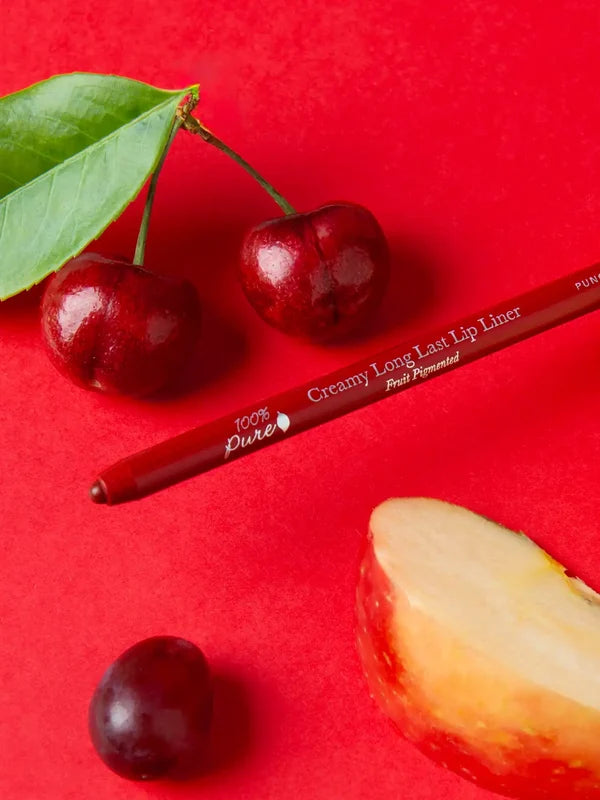 A red 100% Pure lip liner lies diagonally on a bright red surface beside cherries, a green leaf, a dark grape and a pale fruit wedge with red skin. Its exposed tip points toward the lower left.