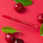 A red 100% Pure lip liner lies diagonally among glossy red cherries and green leaves on a red surface. The exposed tip points left and Shirley Temple is printed near the other end.