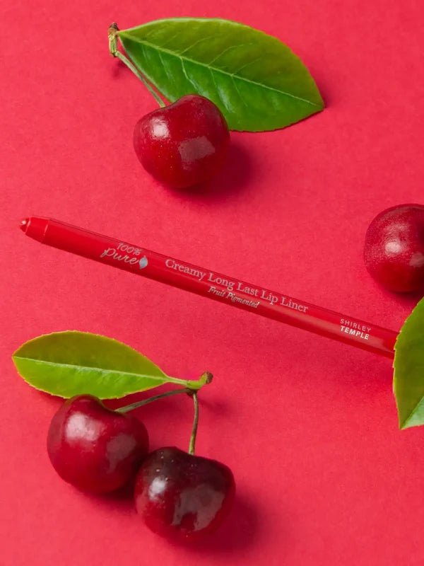 A red 100% Pure lip liner lies diagonally among glossy red cherries and green leaves on a red surface. The exposed tip points left and Shirley Temple is printed near the other end.