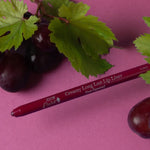 A deep berry-coloured 100% Pure lip liner lies diagonally beside dark grapes and green vine leaves on a purple-pink surface. Its exposed tip points left and the barrel reads Vino.