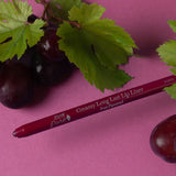 A deep berry-coloured 100% Pure lip liner lies diagonally beside dark grapes and green vine leaves on a purple-pink surface. Its exposed tip points left and the barrel reads Vino.