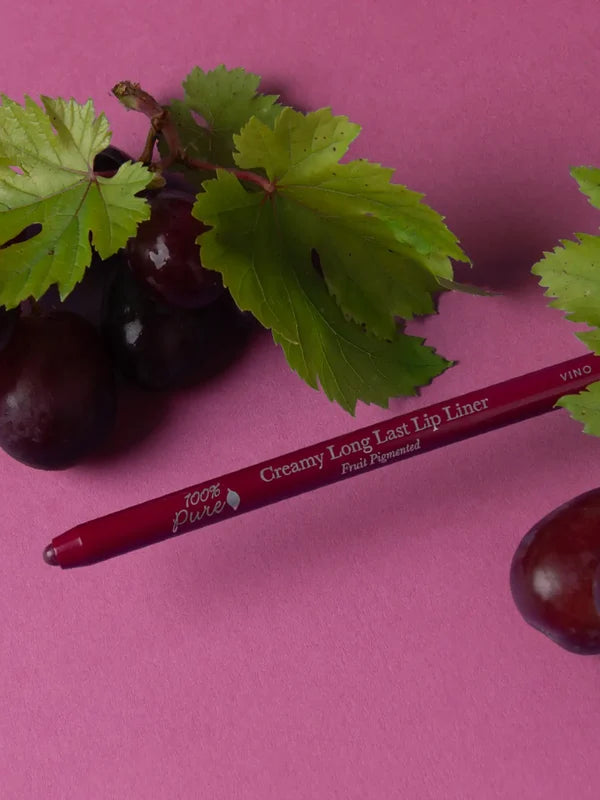 A deep berry-coloured 100% Pure lip liner lies diagonally beside dark grapes and green vine leaves on a purple-pink surface. Its exposed tip points left and the barrel reads Vino.