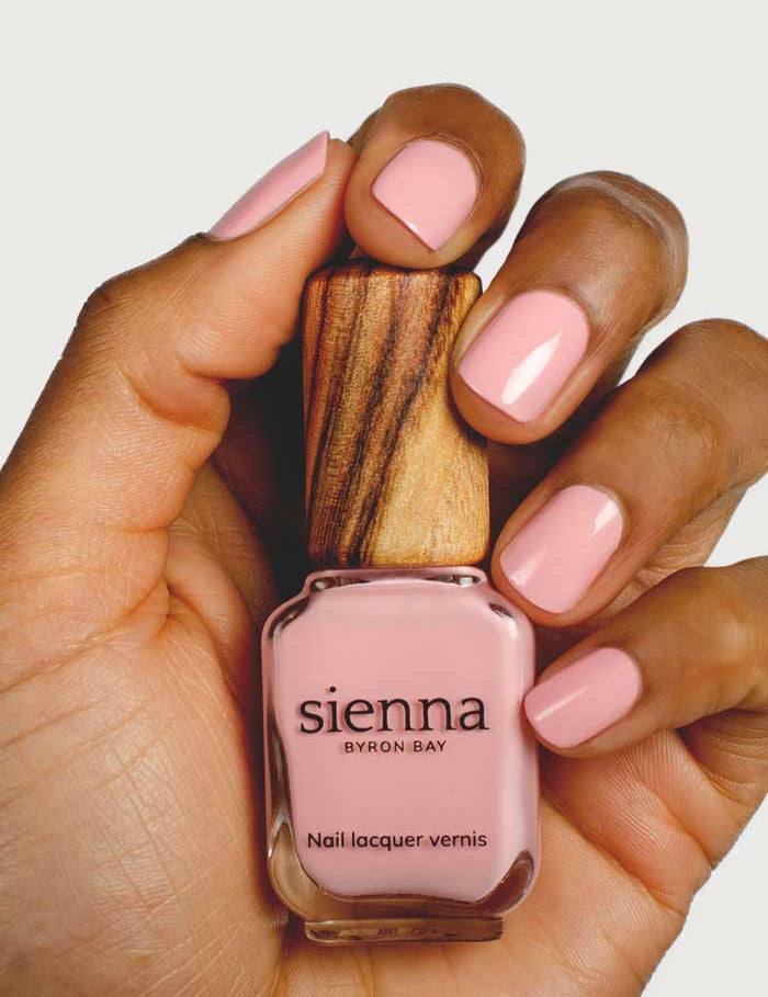 A hand with neatly manicured nails painted in Peony holds a wooden-capped bottle of Sienna Byron Bay Nail Polish - Peony, both in the same plant-derived shade.