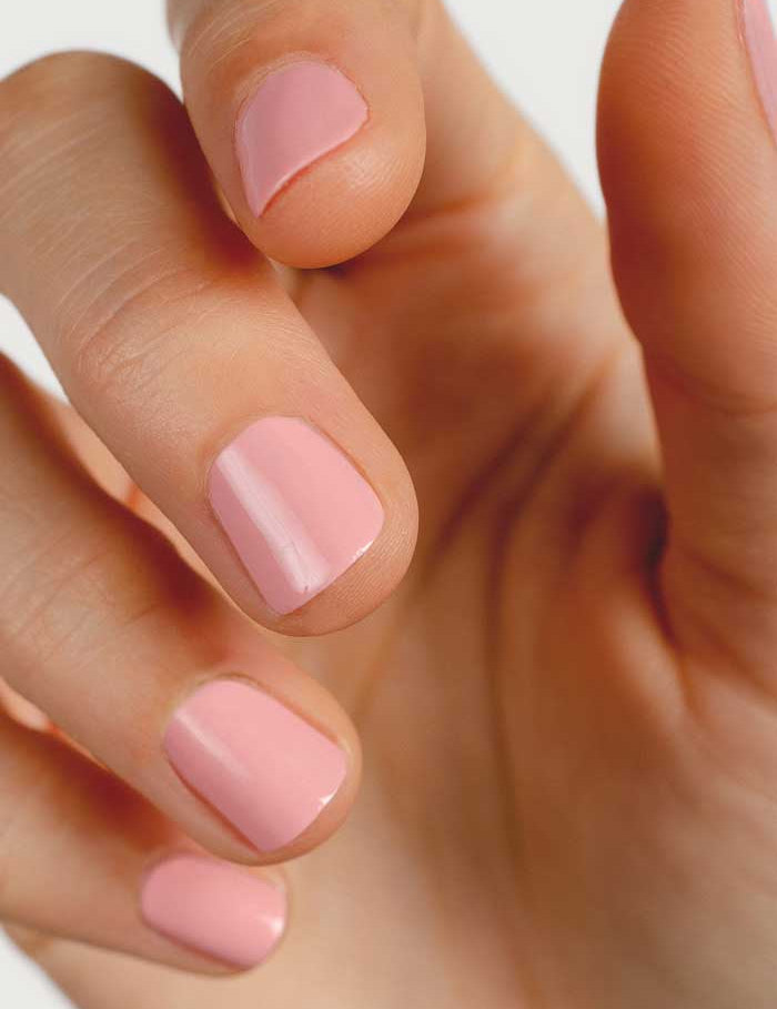 A close-up of a hand with neatly manicured nails painted in Sienna Byron Bay Nail Polish - Peony, a soft cherry blossom pink. The fingers are slightly bent against a plain, light-colored background.