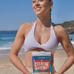 Smiling woman in a pale lilac sports top holding a Botanika Blends Creatine Monohydrate pouch at the beach, with blue sea and sky behind her.