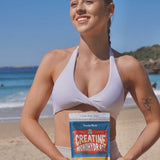 Smiling woman in a pale lilac sports top holding a Botanika Blends Creatine Monohydrate pouch at the beach, with blue sea and sky behind her.