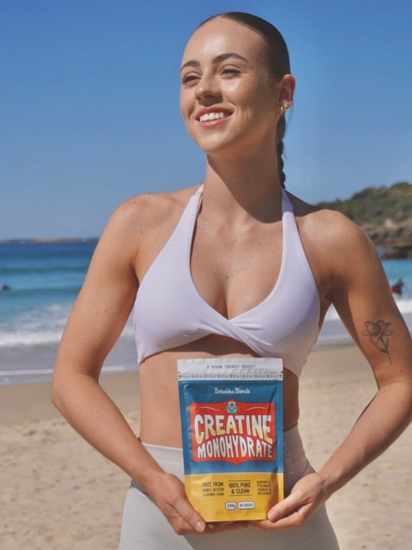 Smiling woman in a pale lilac sports top holding a Botanika Blends Creatine Monohydrate pouch at the beach, with blue sea and sky behind her.