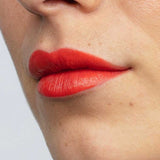 Close-up of lips wearing Antipodes Moisture Boost Natural Lipsticks 4g in Piha Beach Tangerine, a bright orange shade. Smooth skin with small moles is visible; lipstick is enriched with moisturising avocado oil.