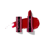 Poppy lipstick in rich red, standing in a reflective pink tube beside its botanical patterned cap, with a matching glossy red smear behind.