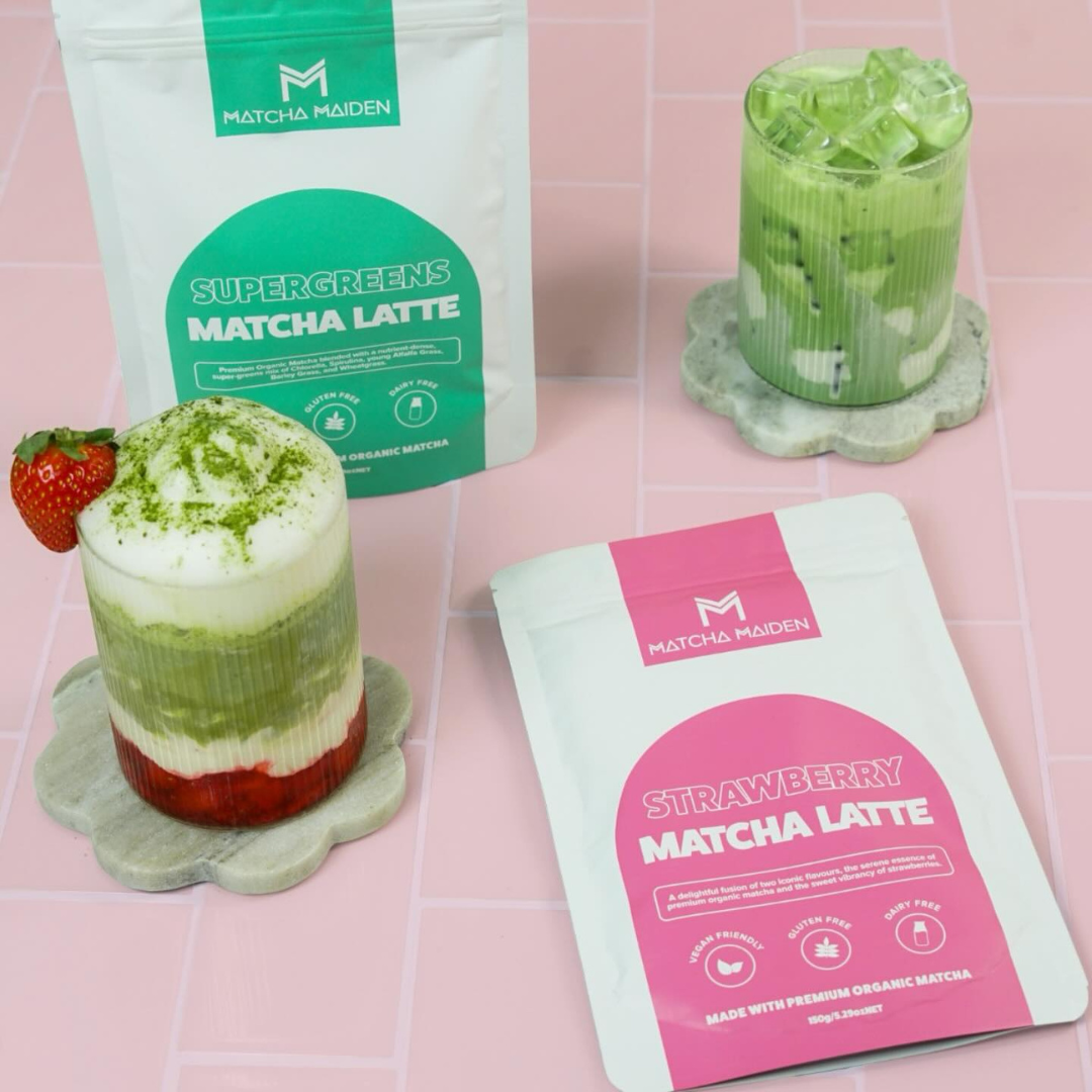 Two glass matcha lattes, one with strawberries and foam, and another with ice, sit on flower-shaped coasters next to Matcha Maiden Supergreens and Strawberry Matcha Latte packets on a pink tiled surface.
