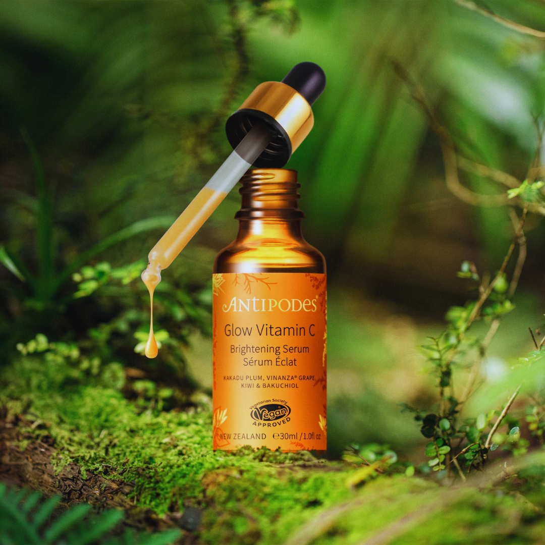 Amber glass bottle of FREE Antipodes Vitamin C Brightening Serum GWP with a dropper, shown dispensing serum over green moss with a lush, blurred forest backdrop.