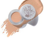 An open jar of 100% Pure Fruit Pigmented® Long Last Concealer with Super Fruits 3g in White Peach, featuring a botanical illustration on the lid, sits atop a swatch of its peachy-beige shade.