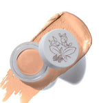 An open container of 100% Pure Fruit Pigmented® Long Last Concealer with Super Fruits 3g rests on a beige smear. The botanical lid lies nearby, and the word “Crème” highlights this long-lasting 100% Pure concealer.