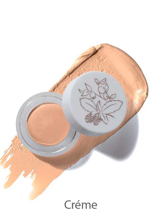 An open jar of 100% Pure Long Last Concealer with a floral lid rests on a beige smear, with the word “Crème” below.