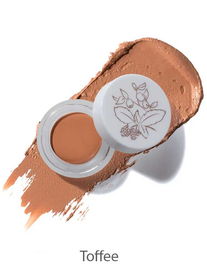An open container of 100% Pure Long Last Concealer 3g in Toffee (medium brown), shown on a textured swatch. The lid displays a botanical illustration.