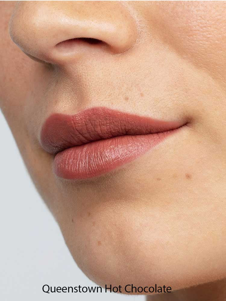 Close-up of smooth lips wearing Antipodes Moisture Boost Natural Lipsticks 4g in a matte mauve-brown shade. The natural formula provides long-lasting color. Text at the bottom reads: Queenstown Hot Chocolate.