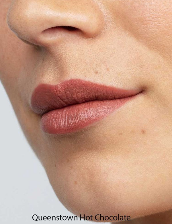 Close-up of smooth lips wearing Antipodes Moisture Boost Natural Lipsticks 4g in a matte mauve-brown shade. The natural formula provides long-lasting color. Text at the bottom reads: Queenstown Hot Chocolate.