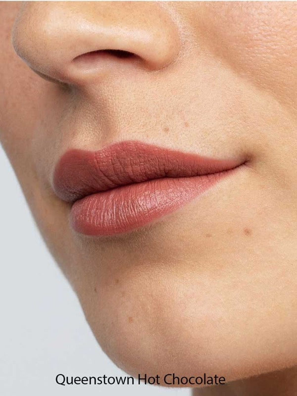 Close-up of smooth lips wearing Antipodes Moisture Boost Natural Lipsticks 4g in a matte mauve-brown shade. The natural formula provides long-lasting color. Text at the bottom reads: Queenstown Hot Chocolate.