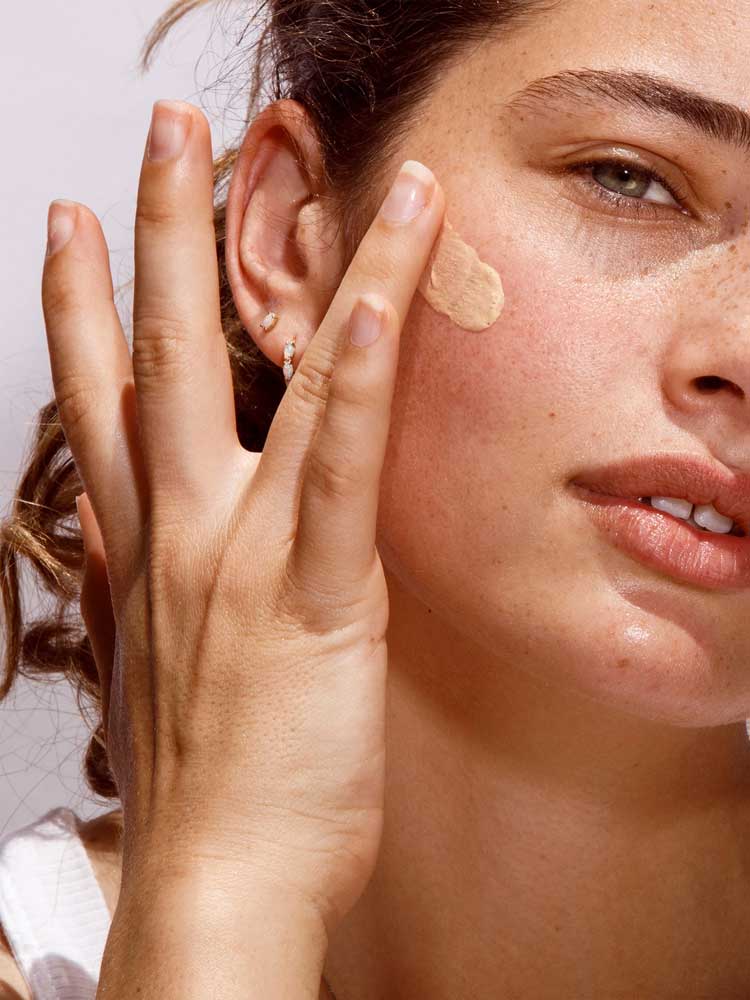 A woman with light skin, green eyes, freckles, and small hoop earrings applies The Organic Skin Co Primp N' Prime Face Primer 60ml to her cheek with her fingers. The well-lit close-up highlights her natural beauty.