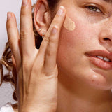 A woman with light skin, green eyes, freckles, and small hoop earrings applies The Organic Skin Co Primp N' Prime Face Primer 60ml to her cheek with her fingers. The well-lit close-up highlights her natural beauty.