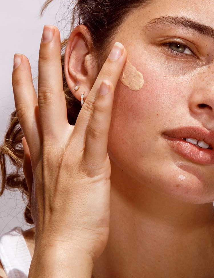 A woman with light skin, green eyes, freckles, and small hoop earrings applies The Organic Skin Co Primp N' Prime Face Primer 60ml to her cheek with her fingers. The well-lit close-up highlights her natural beauty.